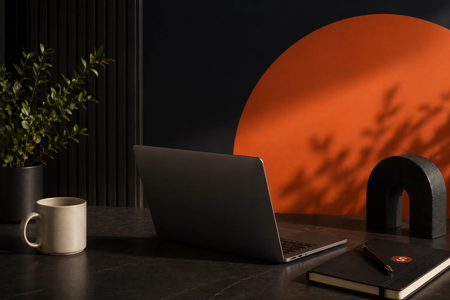 Laptop, notebook, and coffee on a desk against a charcoal and orange wall